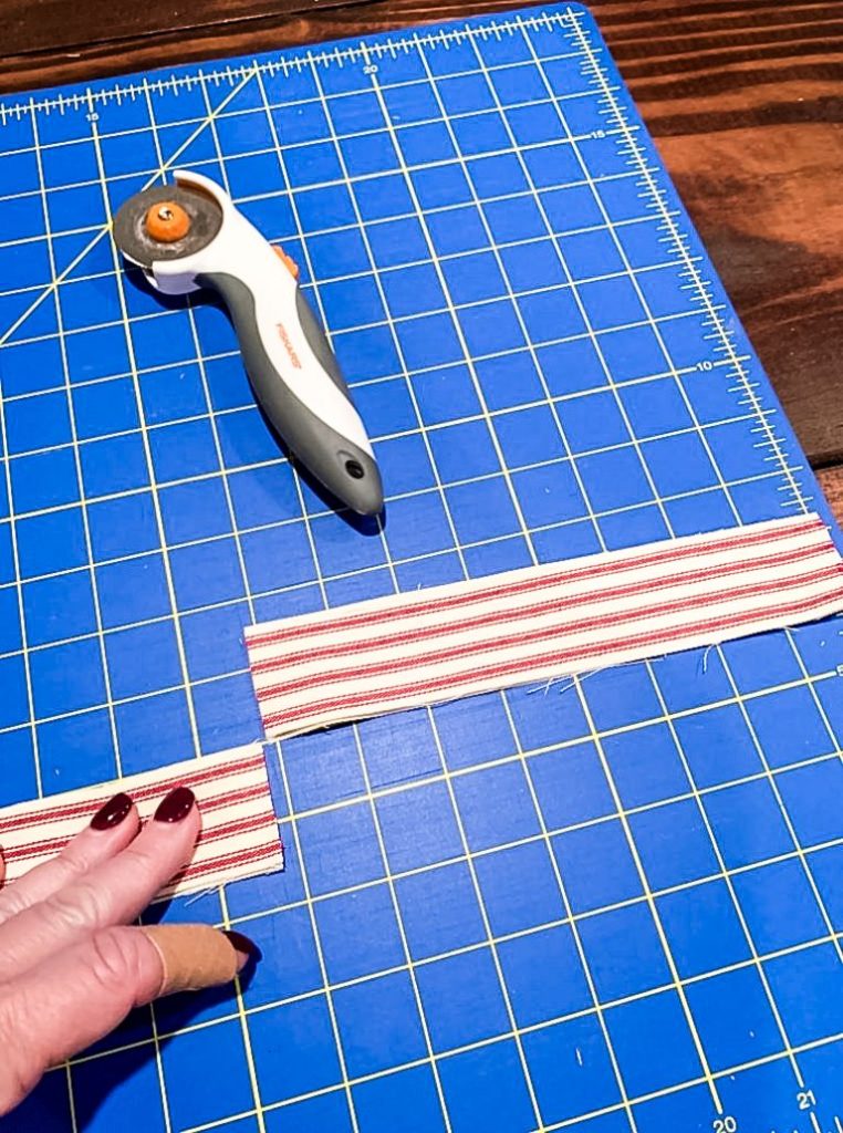 A person's hand cutting a strip of red and white ticking fabric with a rotary cutter on a blue cutting mat.