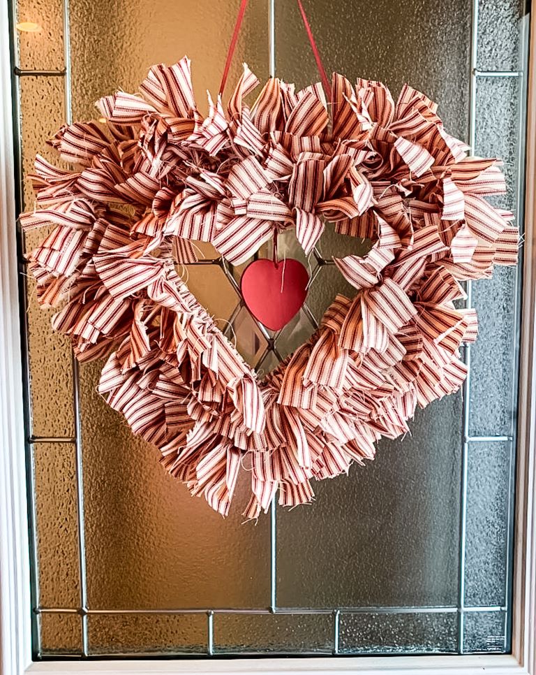 A red and white rag wreath with a small heart accent, hanging on a glass door.
