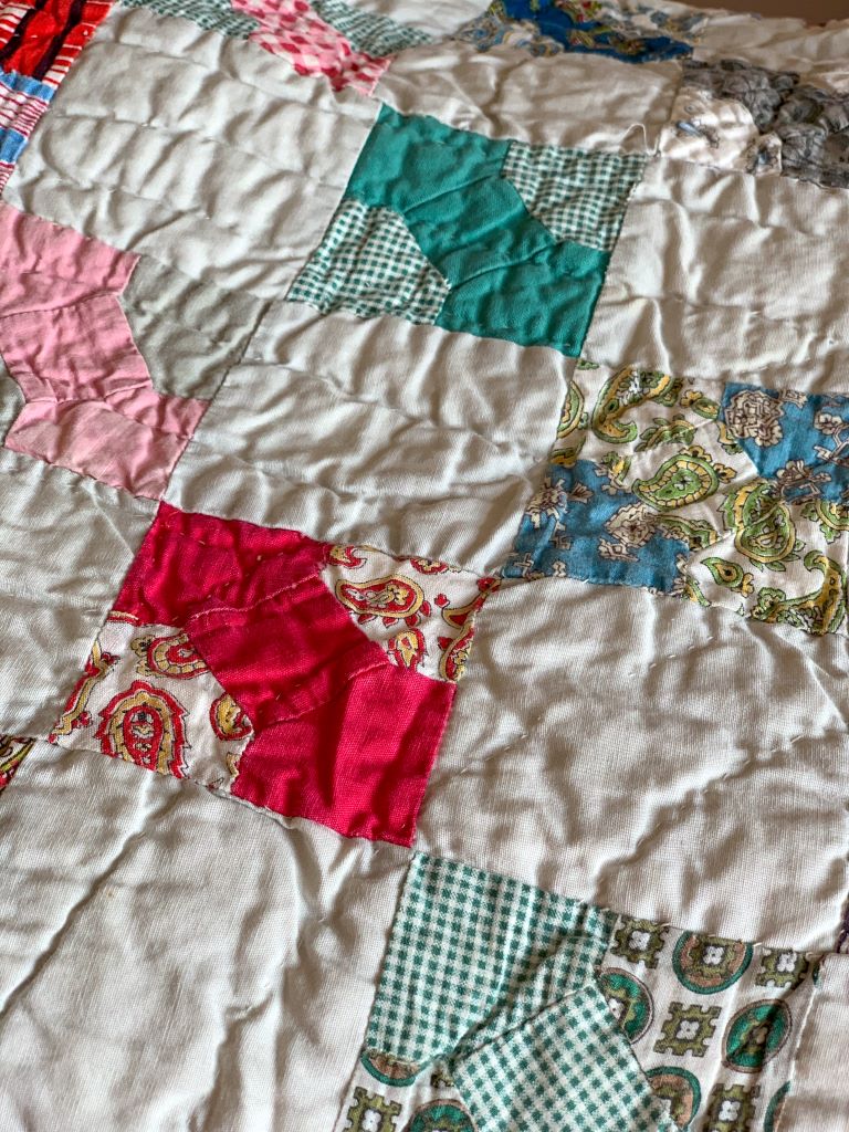Close-up of a vintage handmade patchwork quilt featuring scraps of floral, gingham, and solid fabrics. A traditional cottage heirloom quilt reflecting a storied family history and cozy home decor.