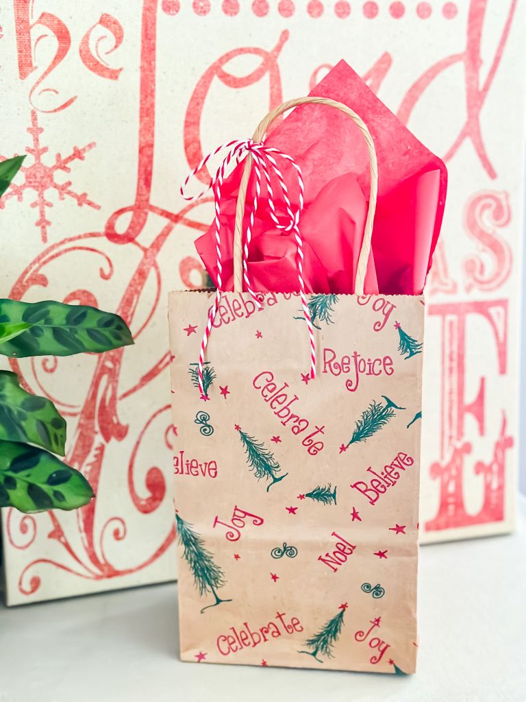 A small brown paper gift bag with handles and a red and white twine bow. The bag is decorated with a repeating pattern of green Christmas trees and red holiday words like 'Celebrate,' 'Joy,' 'Rejoice,' 'Believe,' and 'Noel.' Bright pink tissue paper is visible at the top.