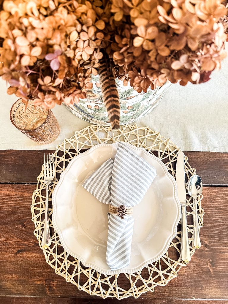 A close-up of a rustic fall tablescape. A white ceramic plate sits on a round straw placemat, topped with a gray-and-white striped linen napkin tied with twine and a small pinecone. A silverware set with a gold detail rests on either side. In the background, there is a vase filled with dried hydrangeas and a single feather, all set on a dark wood table.