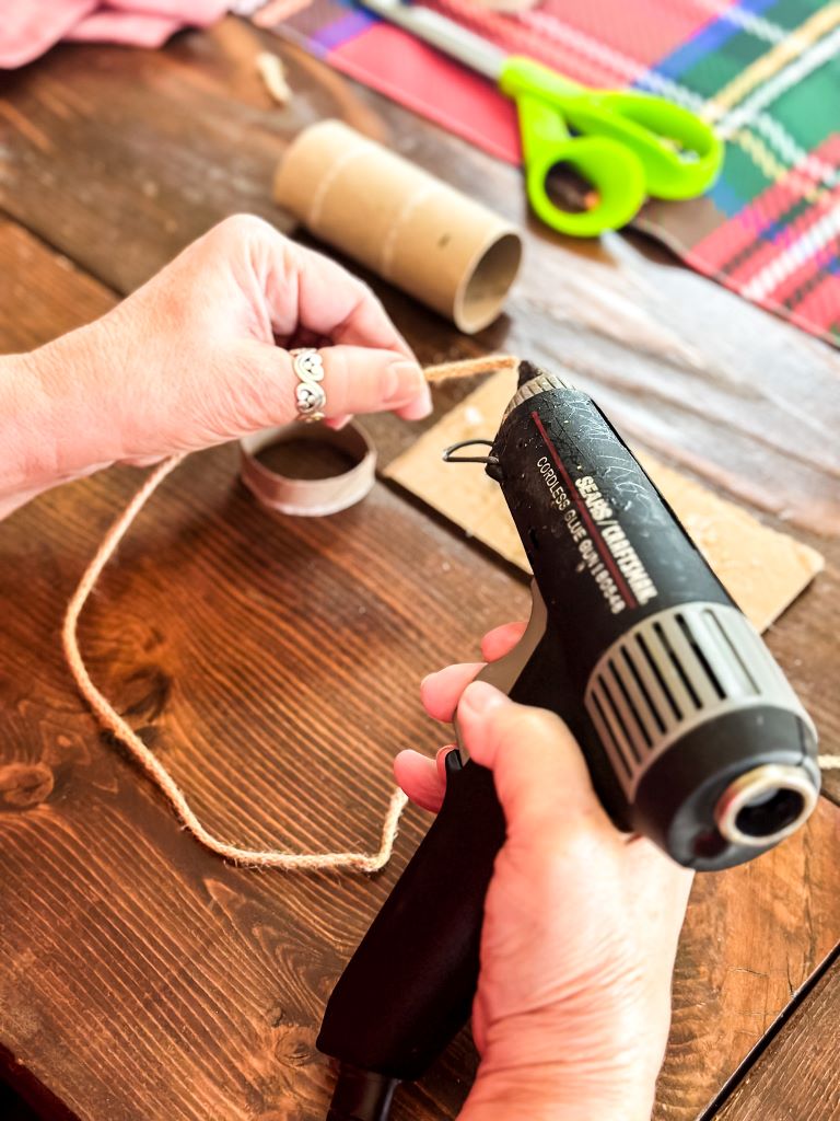 Step-by-step image showing how to use a cordless glue gun to wrap twine around a cardboard base to create a rustic holiday napkin ring for a table setting.
