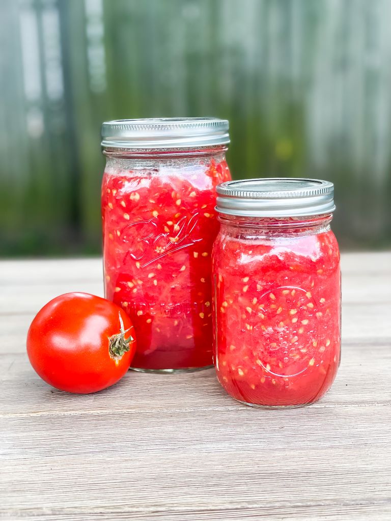 Two glass Ball jars of home-canned crushed tomatoes sitting on a rustic wooden table next to a fresh, ripe red tomato. A perfect example of preserving a summer harvest in a Michigan cottage garden.