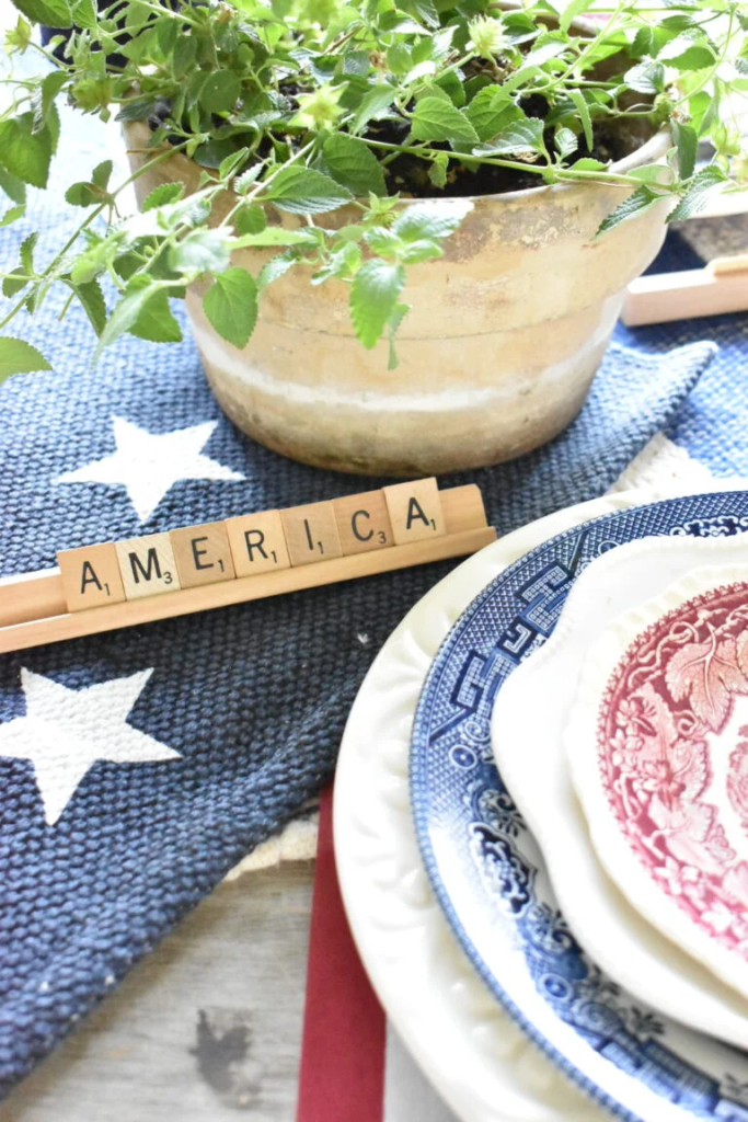 A potted plant on a table with a Scrabble board and plates. The Scrabble board spells out the word "America". Transferware of blue and red are combined with white plates to create a patriotic theme. 