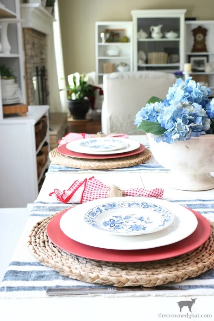 A dining table set with plates, placemats, and a white vessel with blue hydrangeas.