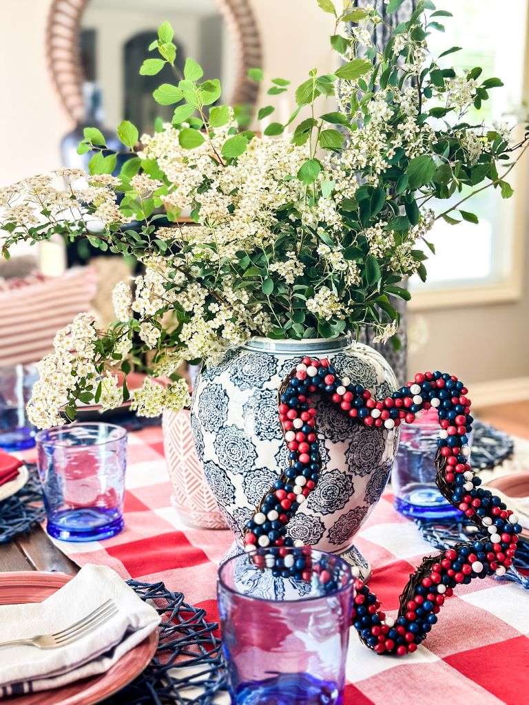 A patriotic table setting with a red and white buffalo check table cloth. A blue and white ginger jar with white flowers as the centerpiece. Red, white and blue table accessories. 