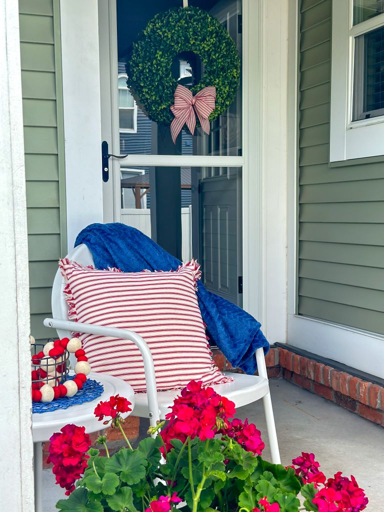 A small side porch with a white table and chair. The chair has a red and white pillow with a navy blue throw. 