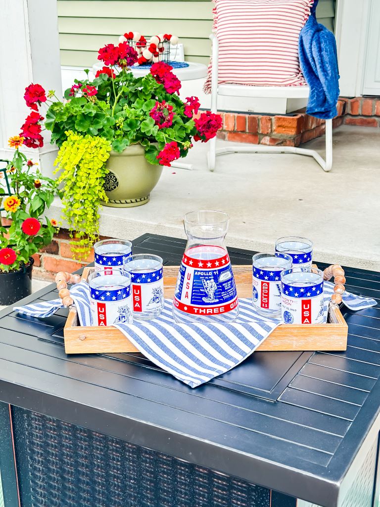 A full image of a wooden tray on a patio table. The tray holds a vintage glass decanter and four vintage glasses with a red, white, and blue Apollo 11 moon landing design. 