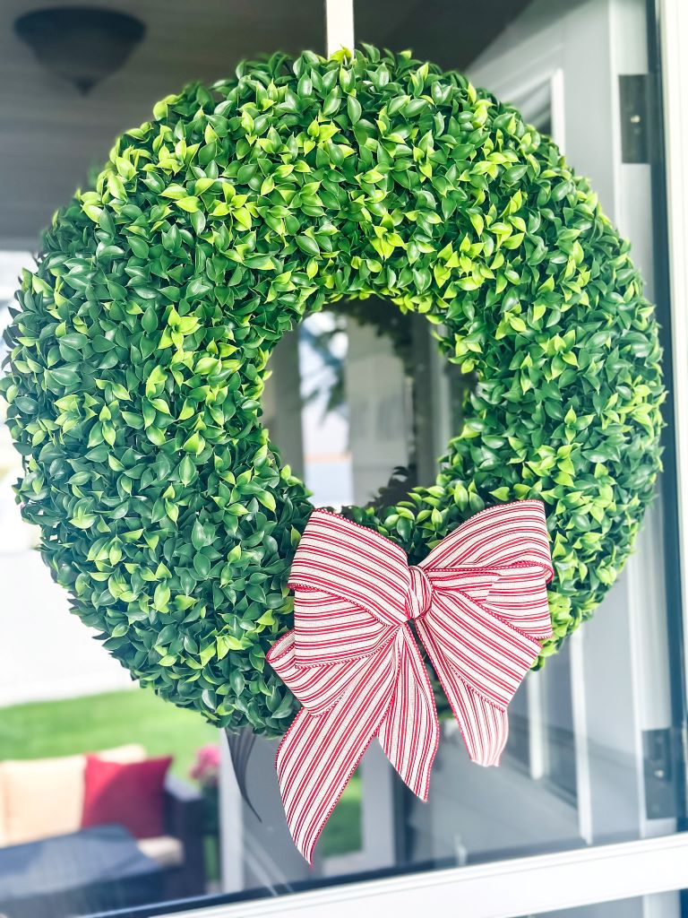 A classic faux boxwood wreath with a simple red and white ticking bow at the bottom. It is hanging on a door. 