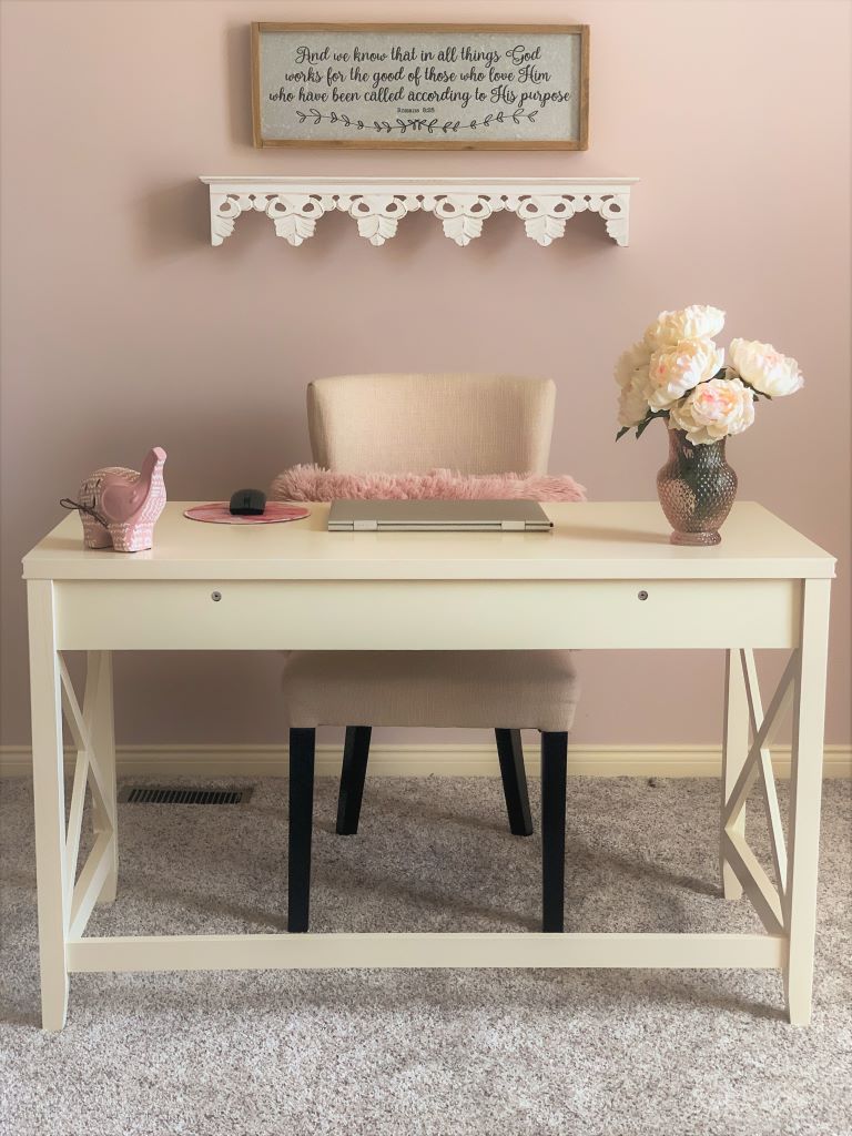  A white desk with a laptop, mousepad, and a pink vase of flowers. A chair with a pink throw is positioned in front of the desk. A shelf with a framed Bible verse hangs above the desk.