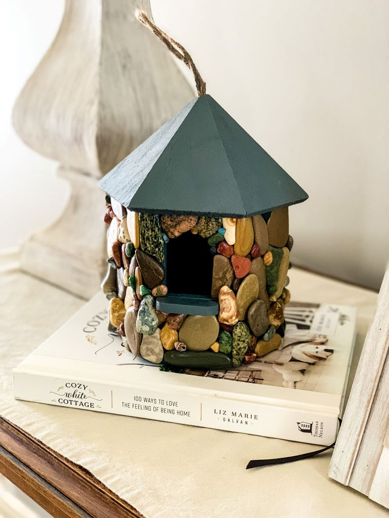 A stone birdhouse with a gray roof and a variety of stones covering the walls, sitting on top of a book titled "Cozy Cottage".