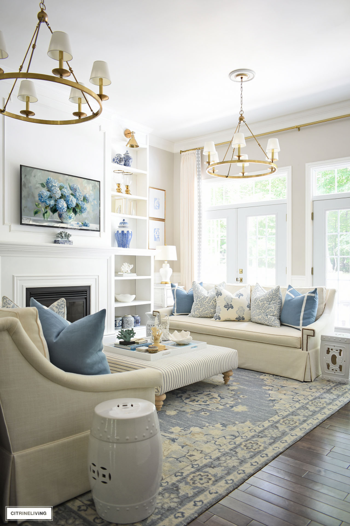  A bright and airy living room with a white sofa and armchair, featuring blue and white patterned pillows. The room has a white fireplace with a blue and white painting above it, and large windows with white curtains.