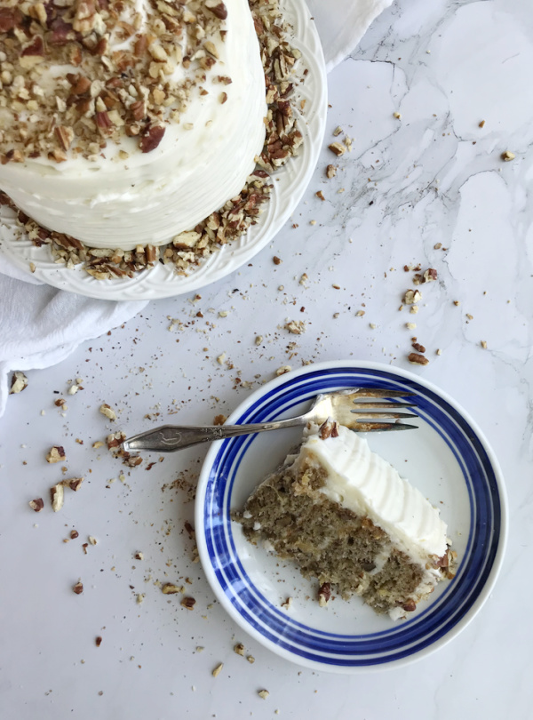 A top-down view of a slice of Hummingbird Cake on a blue and white striped plate, with a fork resting on the plate. The remaining cake is on a white cake stand with a generous layer of cream cheese frosting and chopped pecans.
