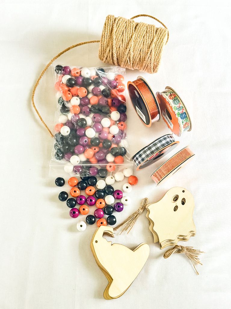 A collection of Halloween crafting supplies for a no-sew no-stress Halloween garland on a white surface. The items include jute twine, colorful beads, ribbons in various patterns, and wooden cutouts of ghosts and witch hats.