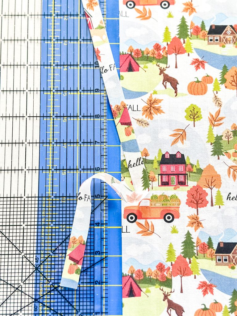 A piece of fabric with a fall-themed pattern being cut on a cutting mat. The fabric features images of a truck, a house, deer, and pumpkins.