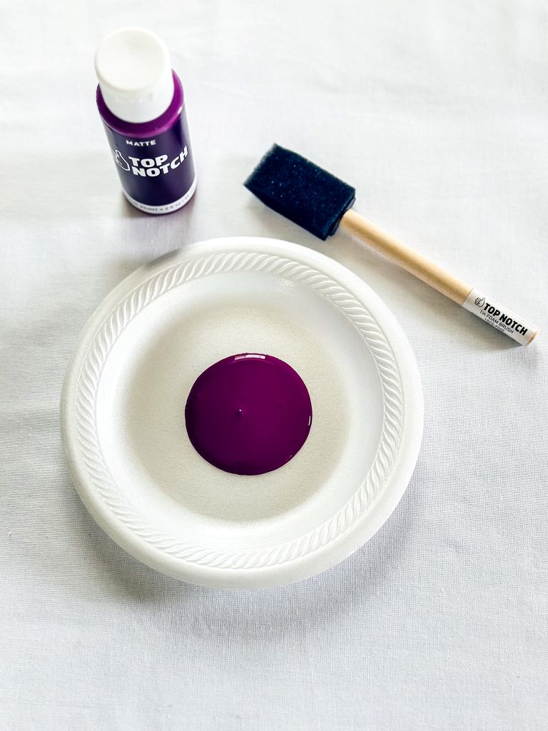 A white paper plate with a dollop of purple paint in the center. A matte paint bottle and a foam brush are also visible on the plate.