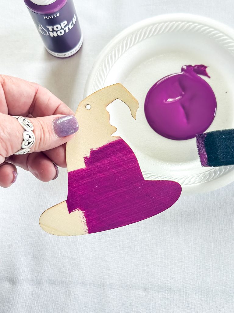 A hand holding a wooden witch hat cutout. The hat is being painted purple with a foam brush. A paper plate with purple paint is also visible in the background.