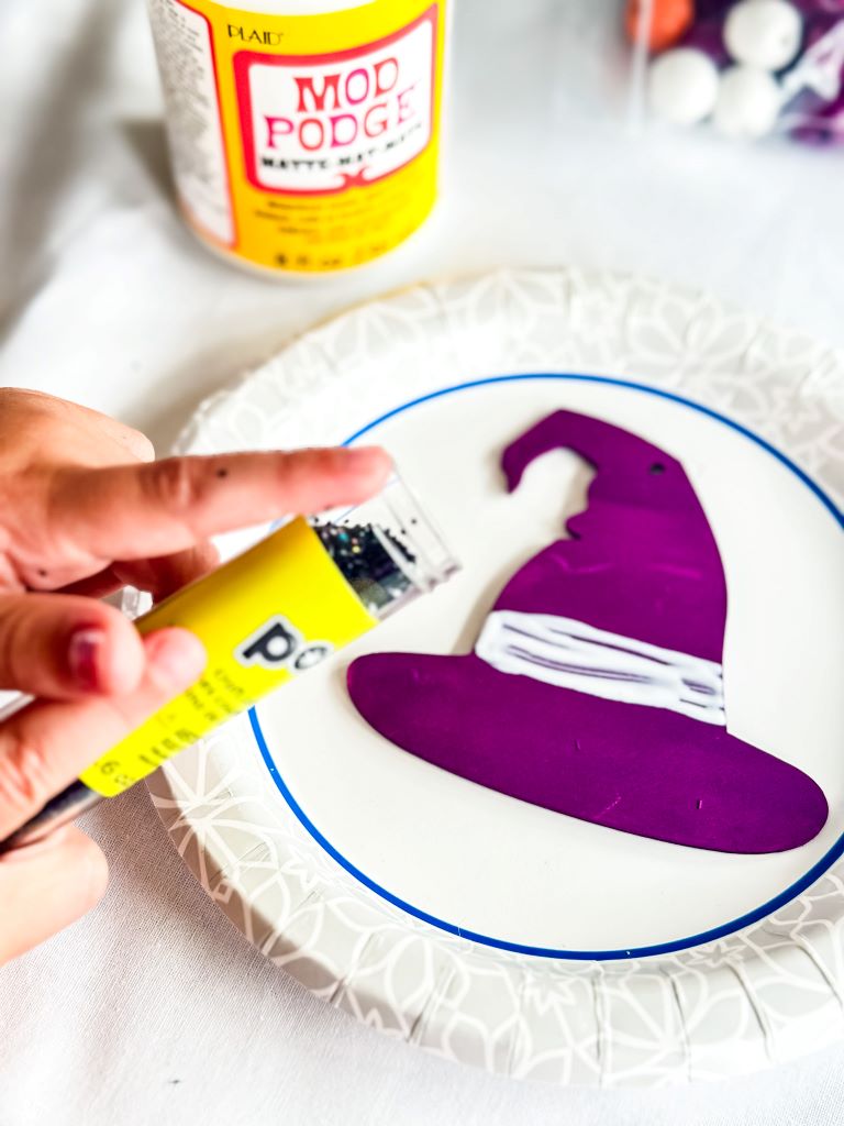 A hand squeezing glitter onto a witch hat cutout that has been coated with Mod Podge. The cutout is resting on a paper plate.
