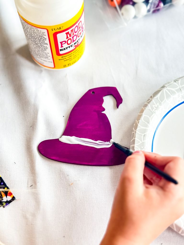 A hand applying Mod Podge with a brush to a purple-painted witch hat cutout. The cutout is resting on a white surface.