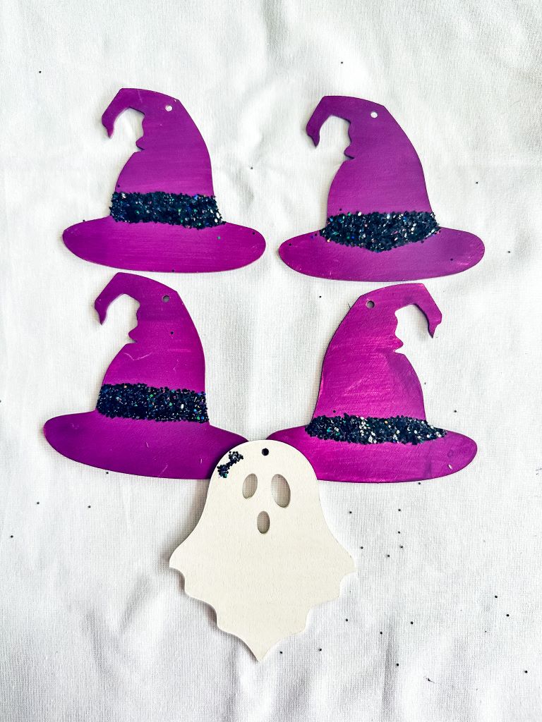 Four purple witch hats and a white ghost cutout arranged on a white surface. The witch hats are decorated with glitter and a black band.