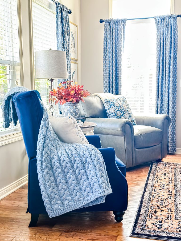 A cozy living room with a blue armchair draped in a chunky knit blanket. A gray leather armchair sits across from it, and a vase of autumnal flowers sits on a small table between them.