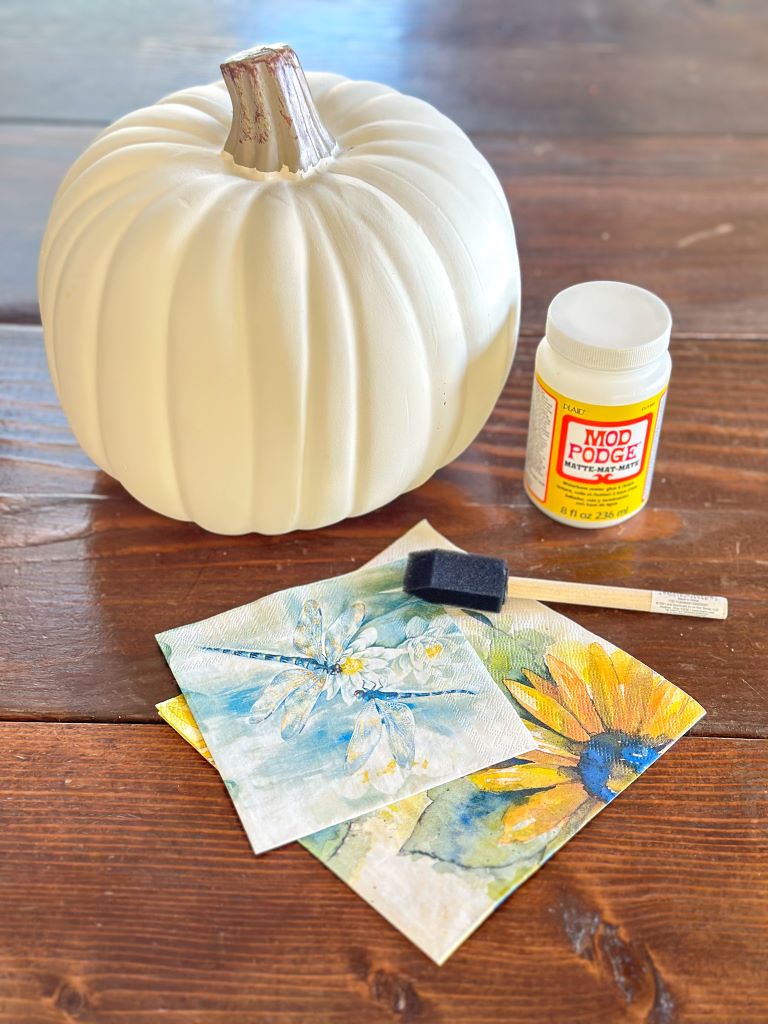 A white craft pumpkin, mod podge, napkins and a small foam brush on a wooden table. 