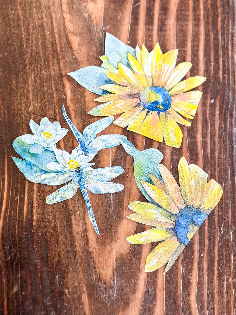 Hand-cut watercolor napkin cutouts of sunflowers, a water lily, and a dragonfly on a wood surface, ready for a decoupage craft project.