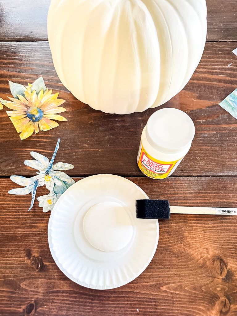A white craft pumpkin sits on a wooden table next to a paper plate covered in white glue. A paintbrush rests on the plate.