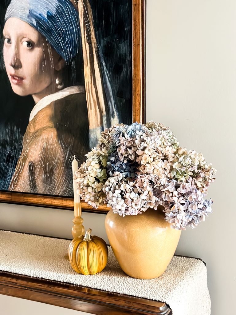 A gold vase of dried hydrangeas on a table with a candle and small pumpkin.
