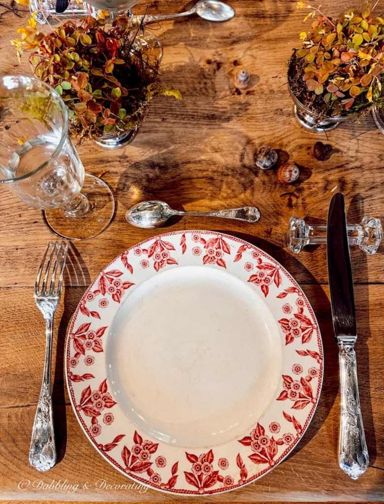 A beautifully set table with a white plate featuring a red floral pattern. Silver cutlery, a crystal knife rest. 