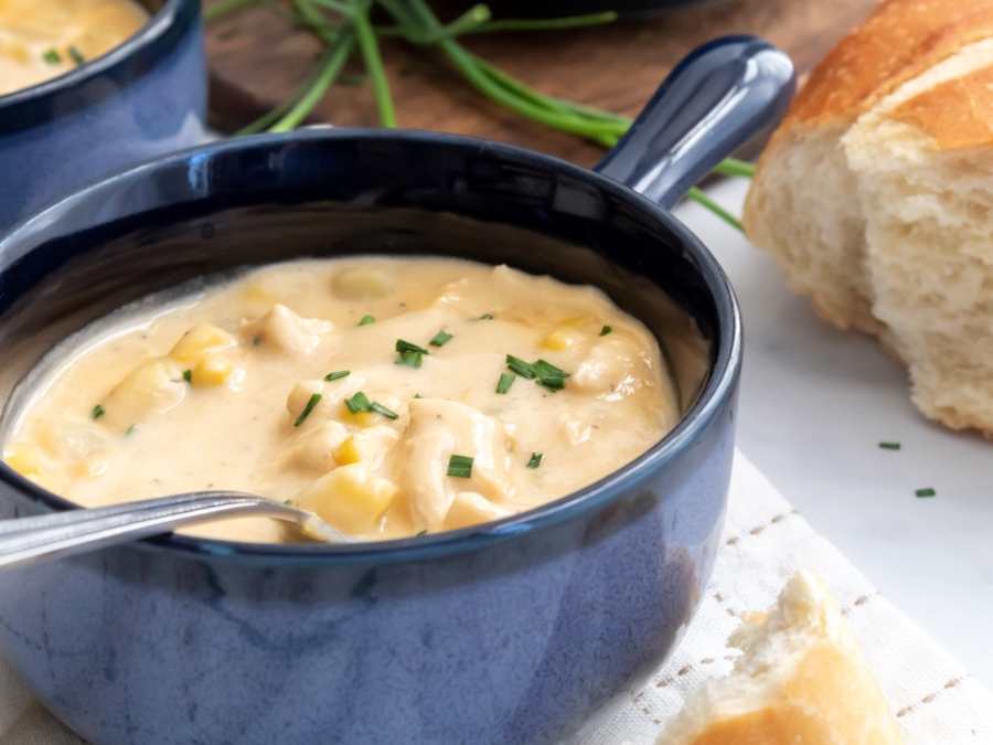 A bowl of creamy chicken and corn chowder, garnished with chopped chives, served with a side of crusty bread.