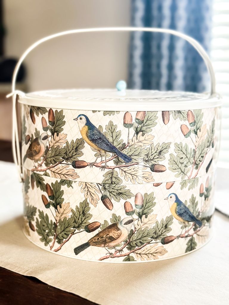 A double decker pie tin with a cream colored background, hosting birds acorns and leaves on it. 