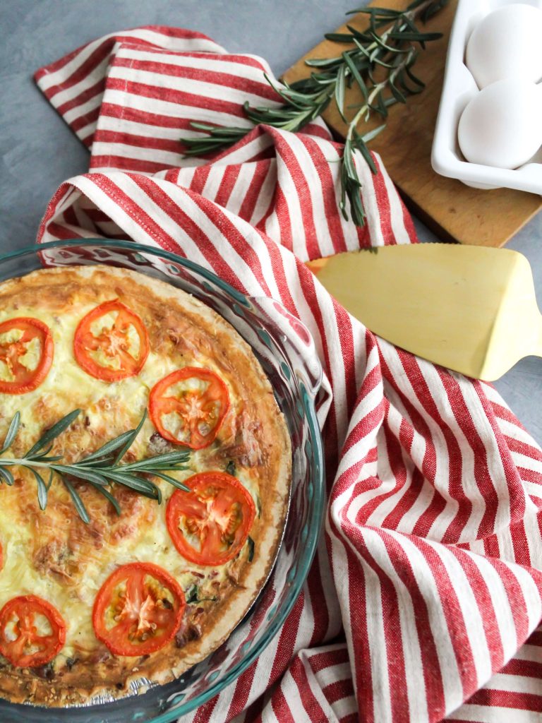 A Christmas quiche topped with tomatoes on a table with a red and white napkin and eggs on the side.