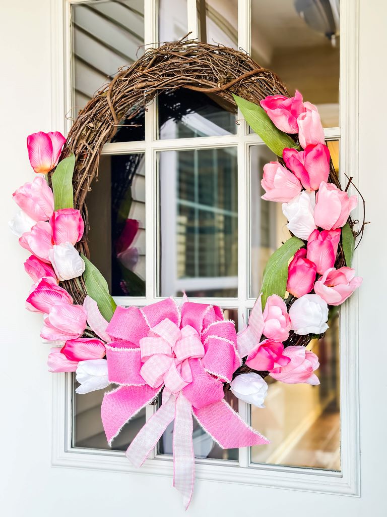 A grapevine wreath with a variation of pink tulips and a large pink and white bow hanging on a door. 