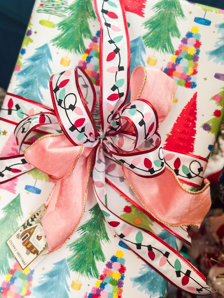 A Christmas gift wrapped in white paper featuring a repeating pattern of colorful, stylized Christmas trees. The present is tied with a bow made of two ribbons: a wide, soft pink satin ribbon and a narrower white ribbon decorated with a string of colorful Christmas lights.