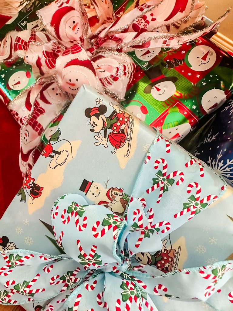 Close-up of a stack of Christmas gifts wrapped in various papers, with the largest in the foreground wrapped in light blue paper. This blue paper features a vintage-style print of Minnie and Mickey Mouse sledding and surrounded by snowmen. The gift is topped with a festive bow made from a light blue ribbon with a repeating pattern of red and white candy canes.