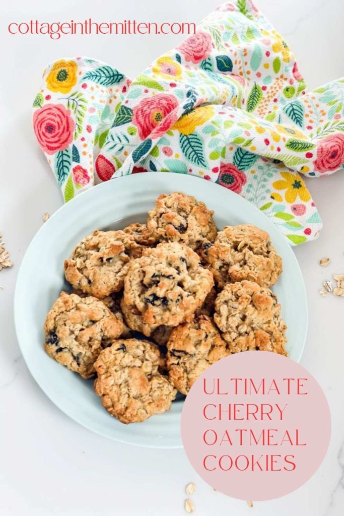 A vertical Pinterest pin for Ultimate Cherry Oatmeal Cookies, featuring a light blue plate piled with chewy oatmeal and dried cherry cookies. The plate sits on a vibrant floral kitchen towel with cottage-style patterns against a clean white background.