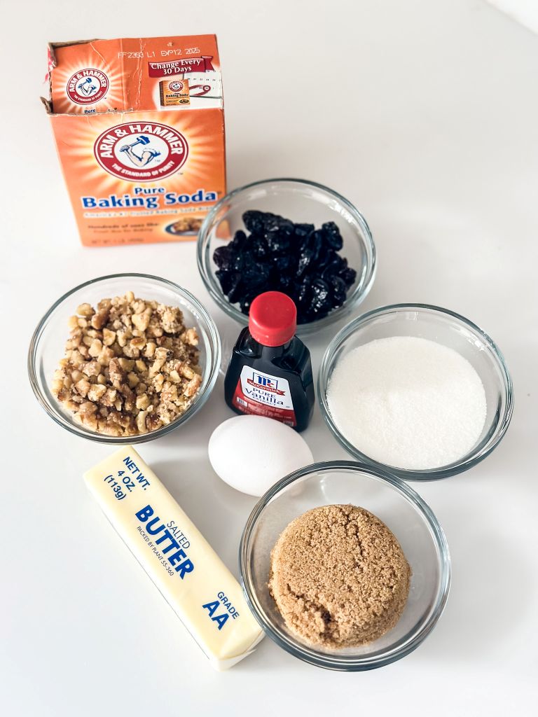 Cookie baking ingredients including butter, brown sugar, white sugar, an egg, vanilla extract, walnuts, dried cherries, and baking soda, arranged in small bowls and containers on a white surface.