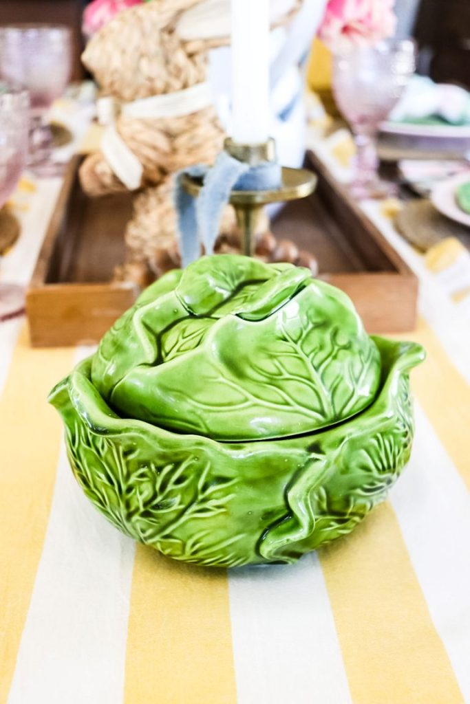 A cabbage pottery piece sitting on a white and yellow table runner with a cottage-style Easter centerpiece in the background. 