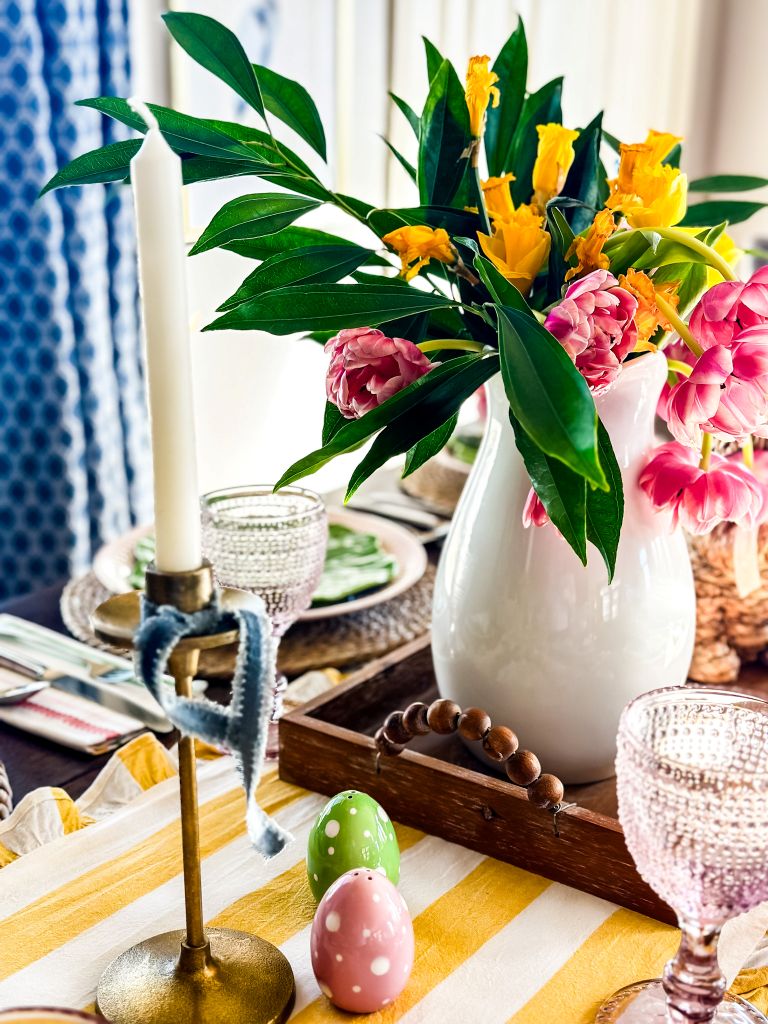 A spring-themed table setting with yellow and white striped runner, pink and yellow tulips in a white vase, a white candle, pink glassware, and painted Easter egg shakers.