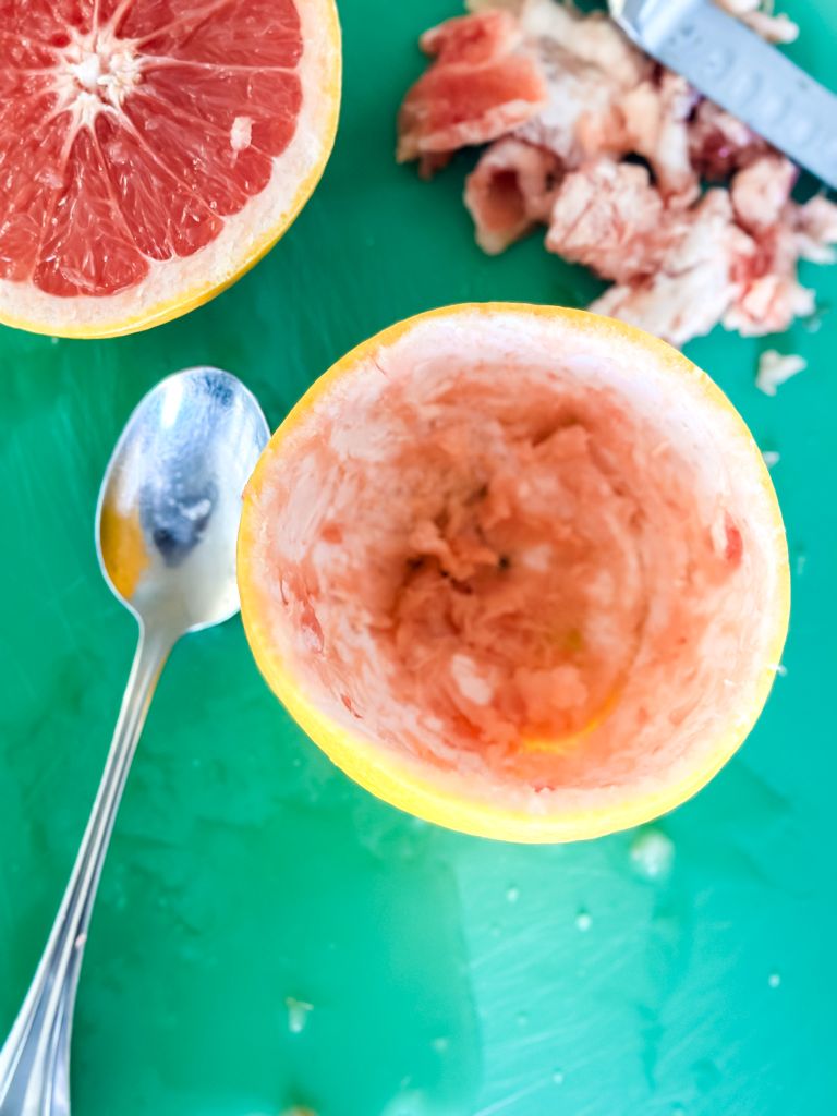 Hollowed-out grapefruit half with a spoon and leftover grapefruit pieces on a green cutting board.