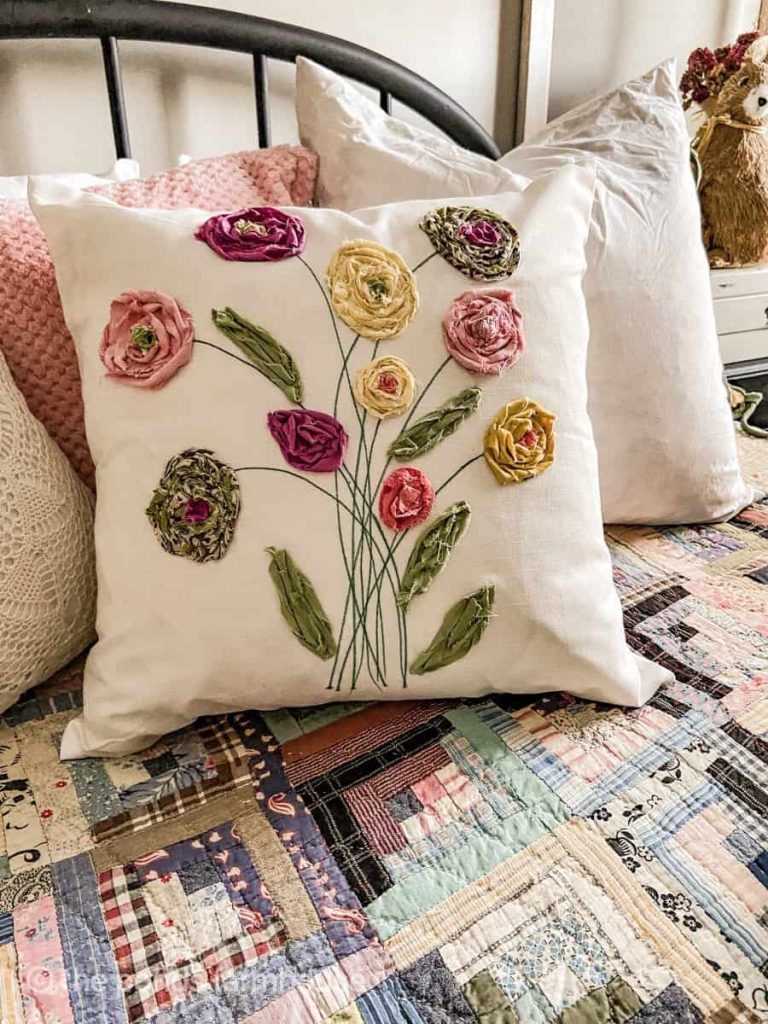 Handmade white pillow with fabric flower appliques in pink, yellow, and green, displayed on a bed with a patchwork quilt and other pillows.