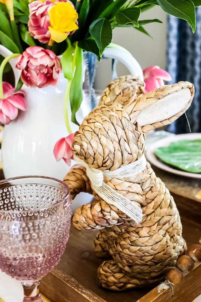 A woven bunny sitting next to a centerpiece made out of a white pitcher, daffodils and pink peony tulips which is sitting on a wooden tray. 