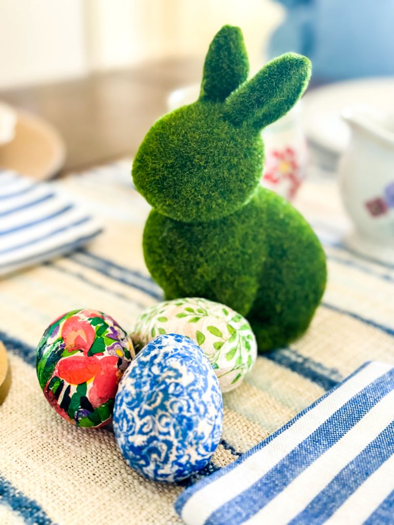 Decoupaged wooden Easter eggs sitting next to a moss bunny on a decorated table for the holiday. 