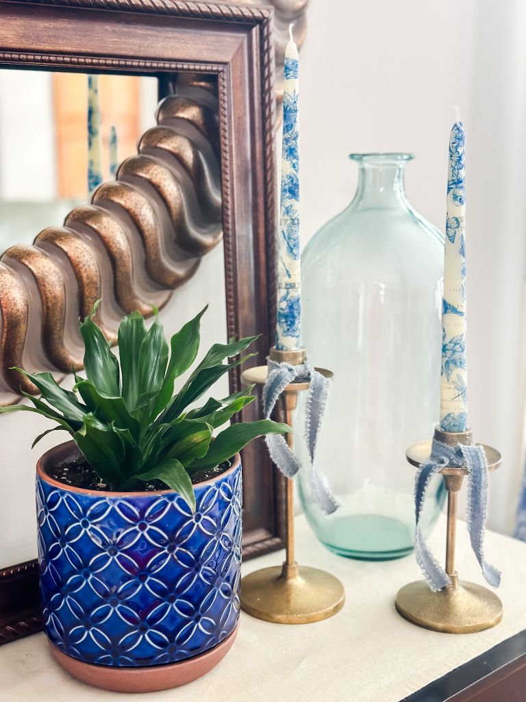 Two taper candles with blue and white patterned napkins ironed on, displayed on brass candle holders with blue ribbon, alongside a potted plant and a decorative mirror.