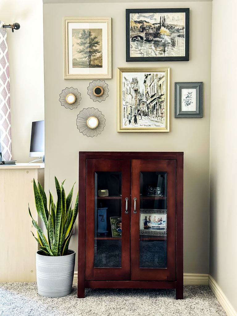 Dark wood cabinet with two glass doors, filled with decorative items, positioned against a beige wall with a gallery wall of framed art. A snake plant in a gray textured pot sits to the left of the cabinet on a light-colored carpet.