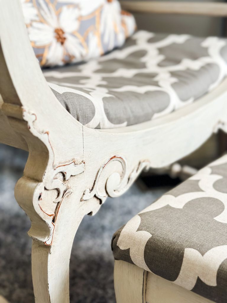 Ornate details on a vintage French chair sitting next to an ottoman upholstered in a grey and beige fabric. 
