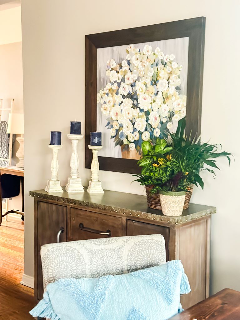A woven basket containing a plant with a mix of green and yellow foliage, positioned on a rustic wood cabinet. Above the cabinet hangs a framed painting of white flowers. Three white candlesticks with dark candles are arranged on the cabinet to the left of the plant.