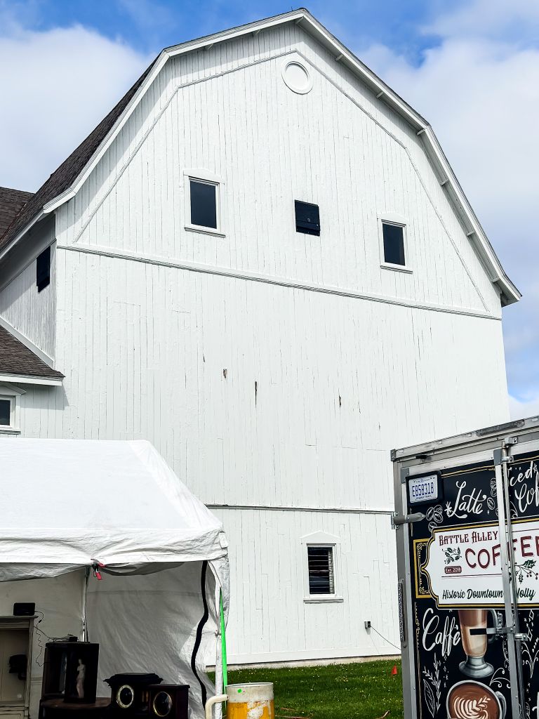 Large white barn with a peaked roof and small dark windows, with a vendor tent and a Battle Alley Coffee sign set up in front on a sunny day.