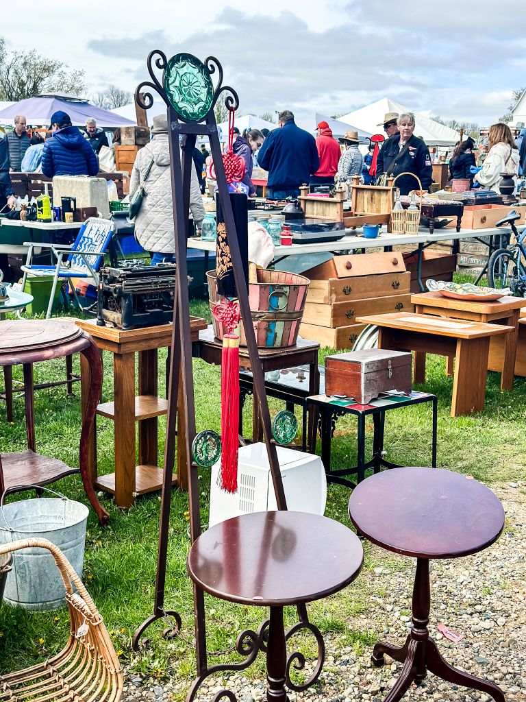 Outdoor vintage market with shoppers browsing rows of tables filled with antique furniture, wooden crates, a vintage typewriter, small side tables, and other treasures under a cloudy sky.